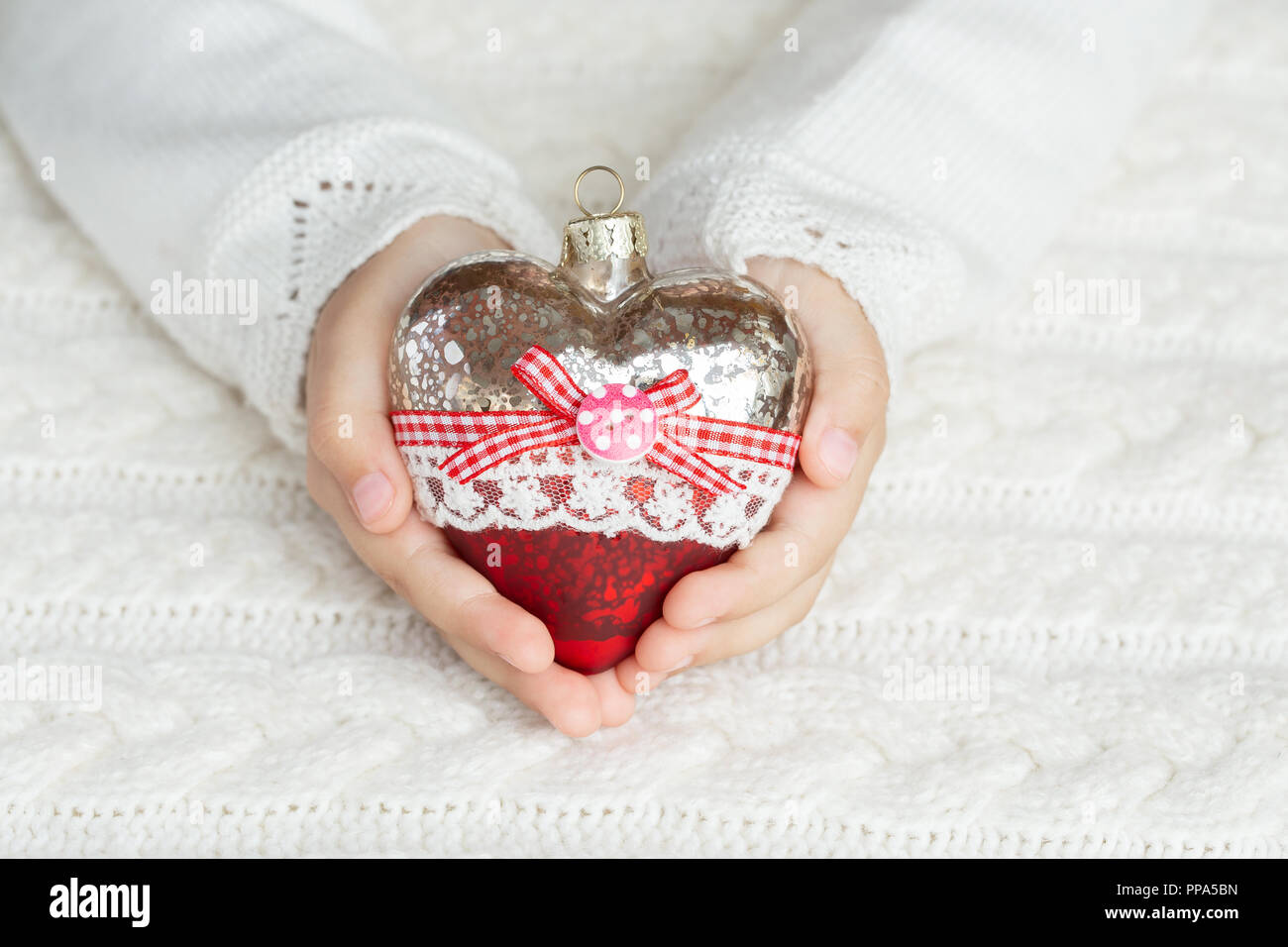 Bambino tiene un cuore di vetro, un giocattolo di Natale nelle mani sullo sfondo di un maglione caldo Foto Stock