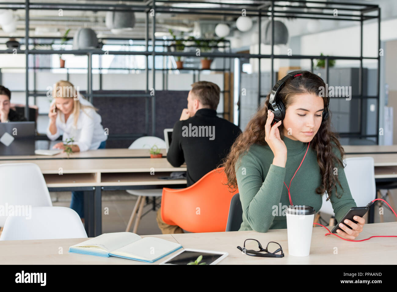 Ritratto di giovane contemporanea donna ascoltare musica al lavoro indossando le cuffie di grandi dimensioni in open space office Foto Stock