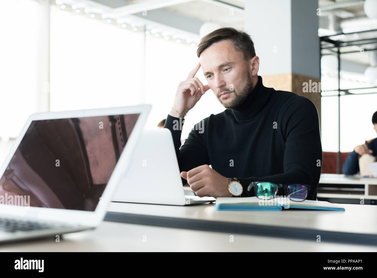 Ritratto di bel giovane guardando lo schermo del notebook pensively mentre si lavora in open space office Foto Stock