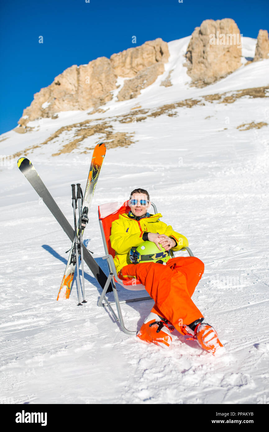 Immagine di sport uomo seduto sulla sedia accanto a sci e bastoni sullo sfondo di montagne innevate Foto Stock