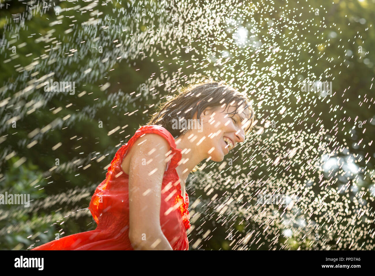 Ridendo donna il raffreddamento sotto un getto di acqua nel suo arancione abiti estivi in un giorno caldo contro uno sfondo di verdi alberi. Foto Stock