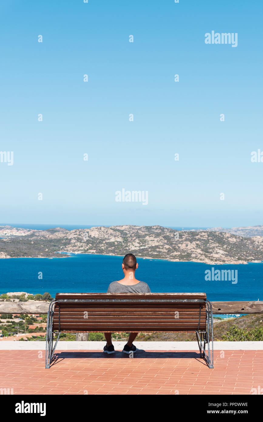 Giovane uomo caucasico, visto da dietro, seduto su una panchina in Palau, nella costa nord della Sardegna, Italia, osservando il mare Mediterraneo e la M Foto Stock