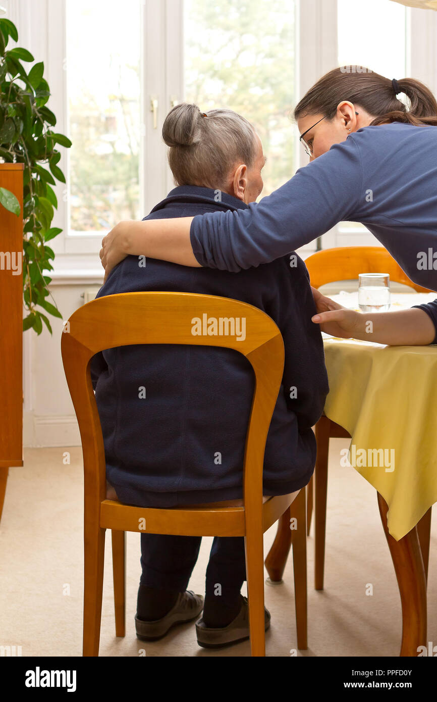 Vista posteriore di un senior donna seduta a casa beeing abbracciato da suo nipote che sta aiutando il suo durante il giorno, eldercare da membri della famiglia Foto Stock