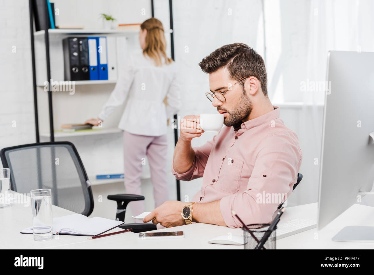 Bel giovane imprenditore di bere il caffè mentre il suo segretario prendendo una cartella dal ripiano sfocati sullo sfondo Foto Stock