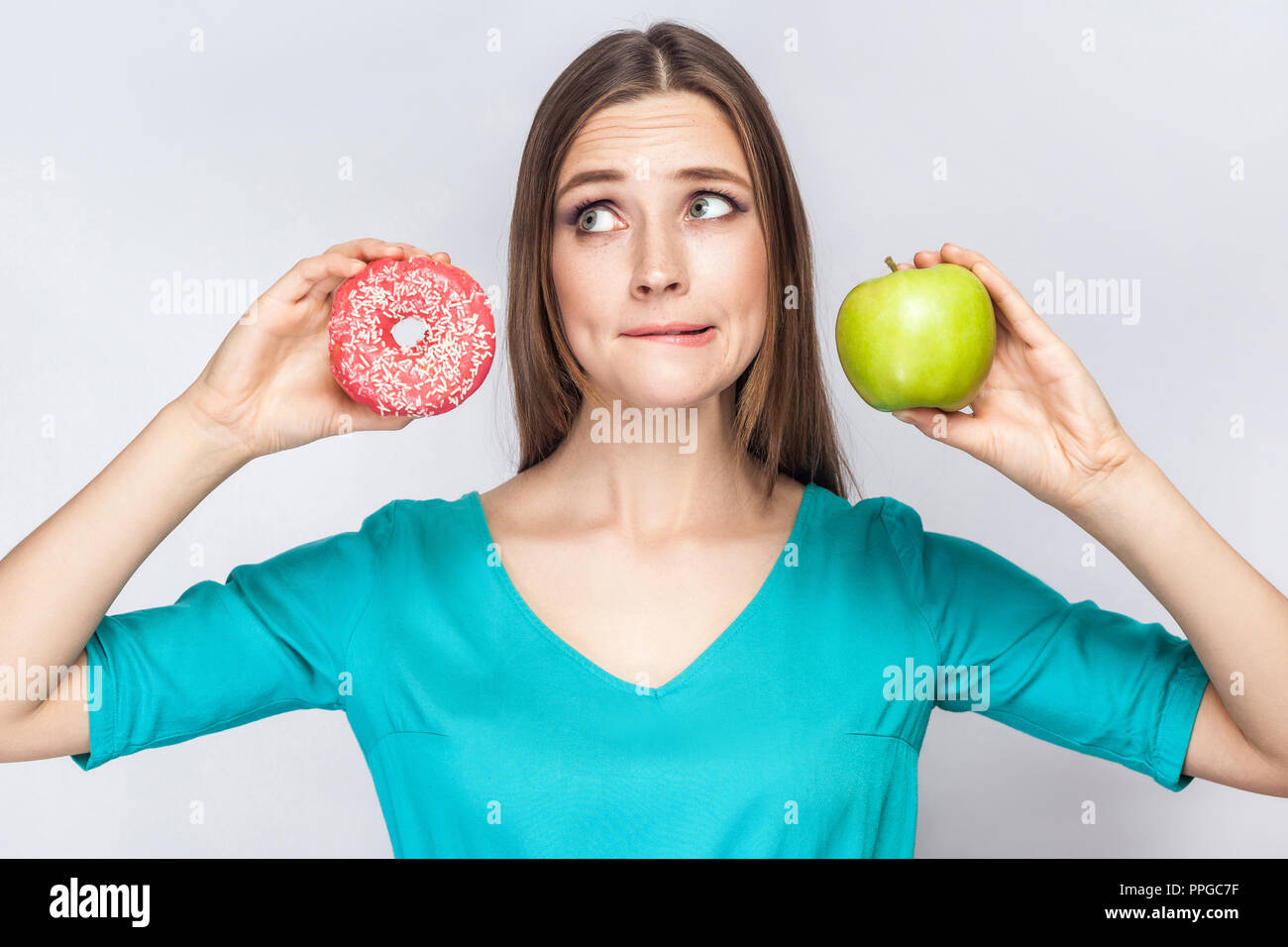 Ritratto di sognare giovane bella ragazza in blu camicetta in piedi, mostrando e azienda ciambella rosa e verde mela in mani con intelligentemente faccia astuto Foto Stock