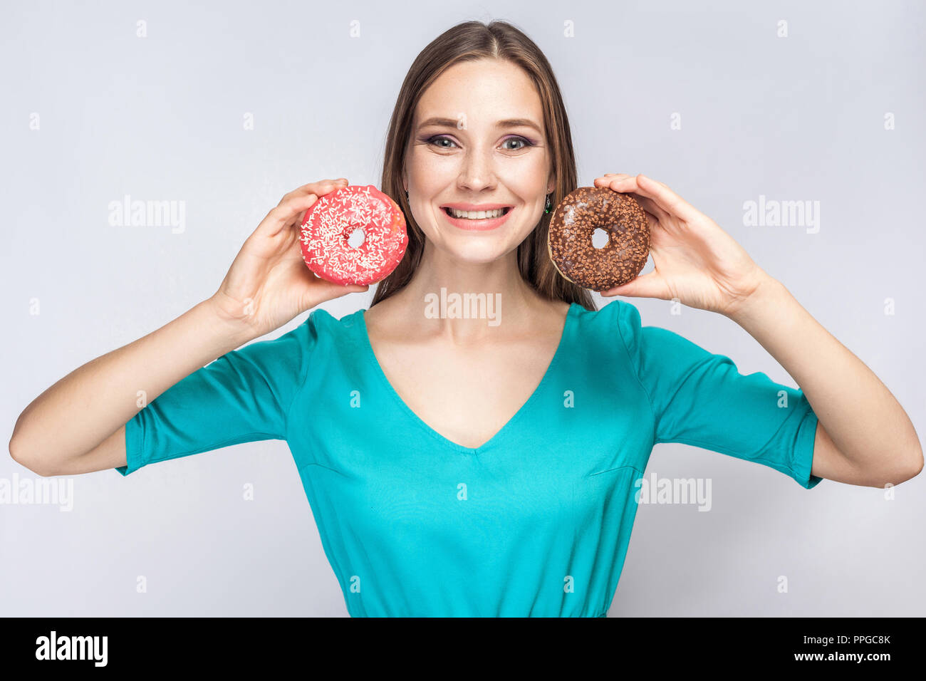 Ritratto di positivo sorridente giovane bella ragazza in blu camicetta in piedi, azienda e mostra rosa e ciambelle al cioccolato con sorriso toothy su grigio bac Foto Stock
