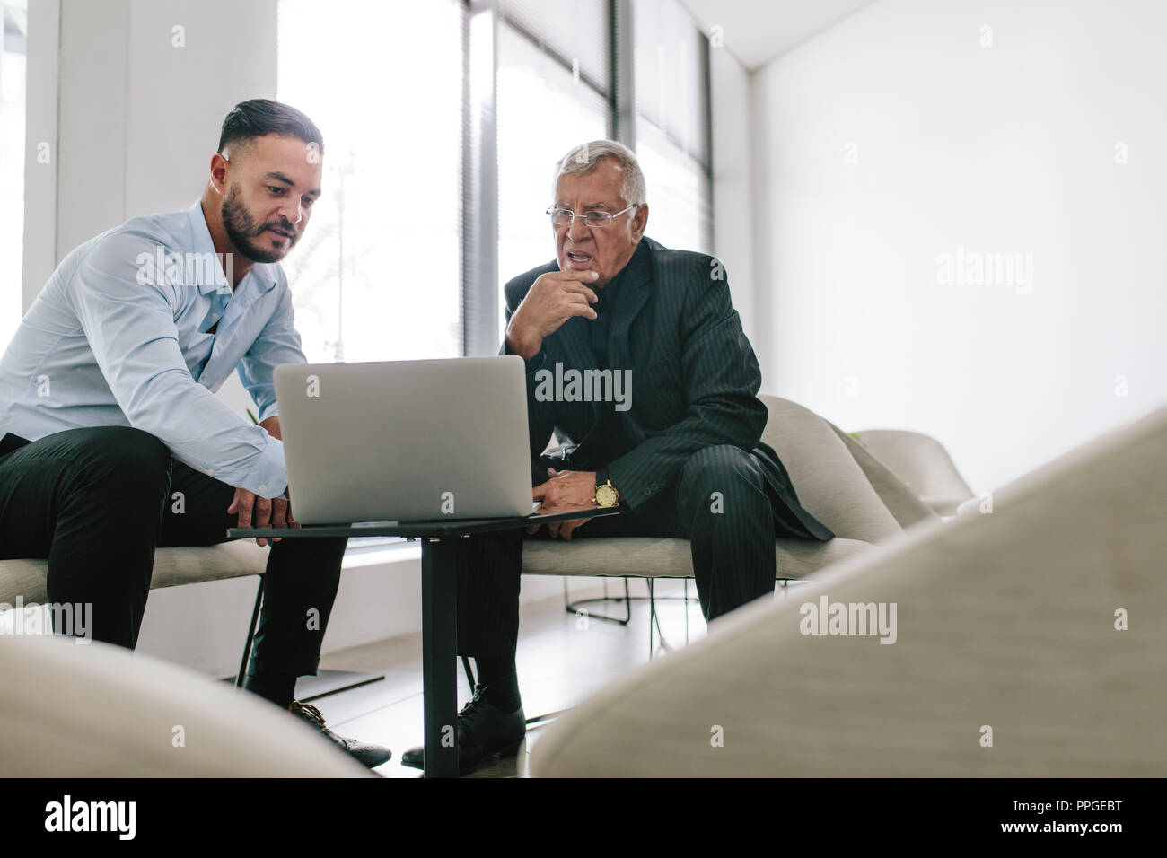 Colpo di due imprenditori utilizzando laptop nella lobby dell ufficio moderno. Giovane dirigente che mostra la sua proposta a un senior uomo seduto nel foyer. Foto Stock