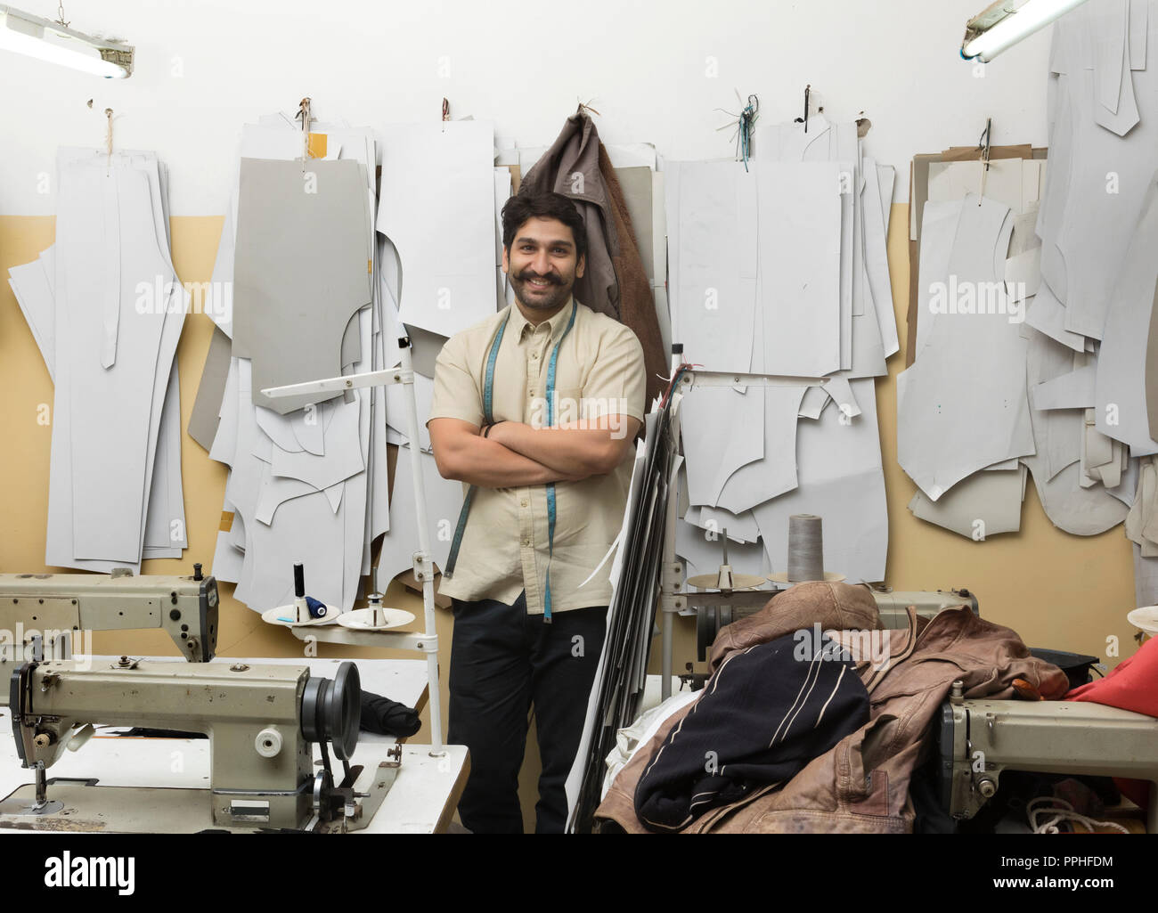Sarto sorridente in piedi con le braccia incrociate nella sua piccola officina con campione di talee appesi in background. Foto Stock