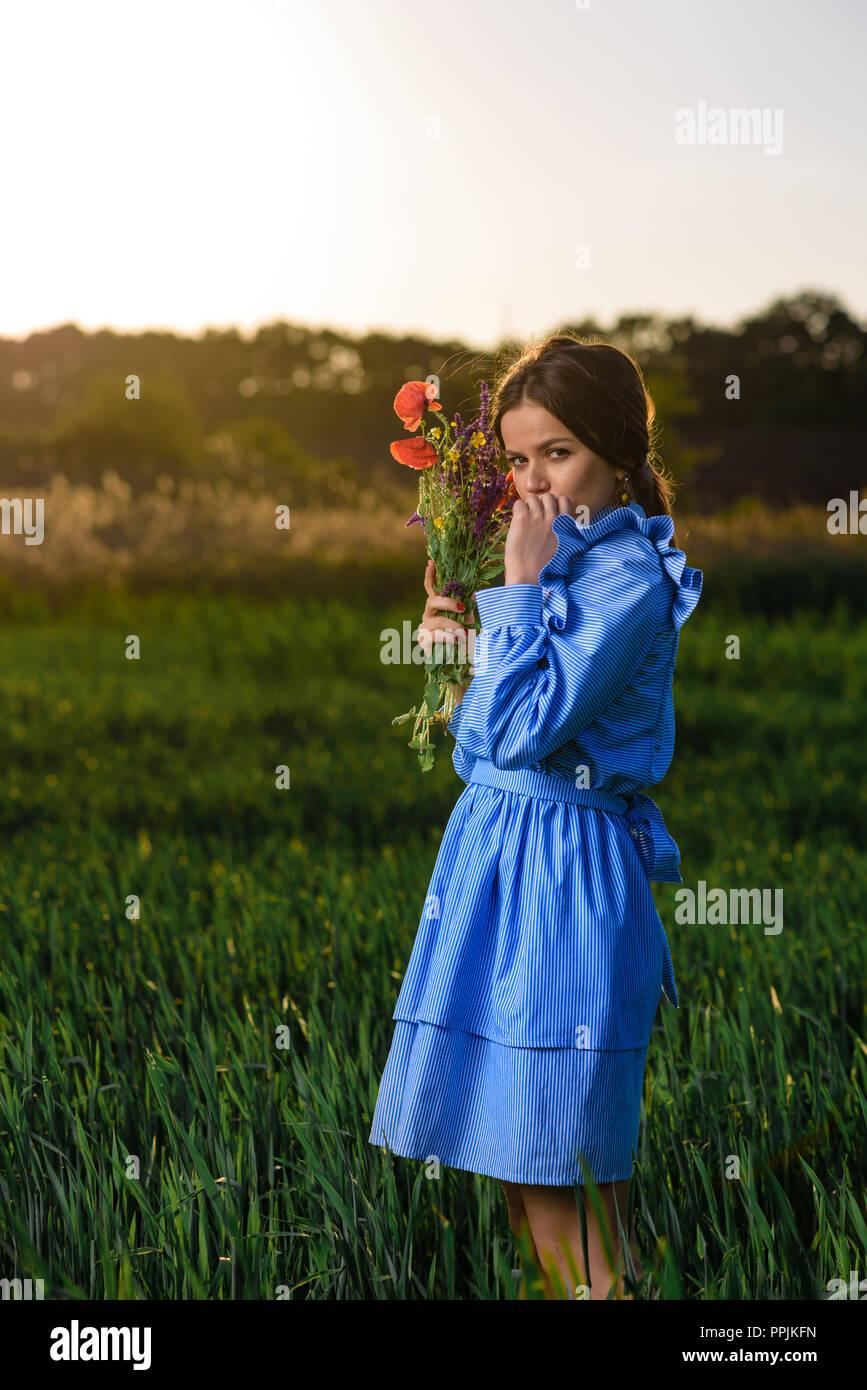 Giovane donna in blu e bianca a strisce abito è in possesso di un bouquet di fiori d'estate e guardando la telecamera mentre in piedi nel campo del verde whea Foto Stock