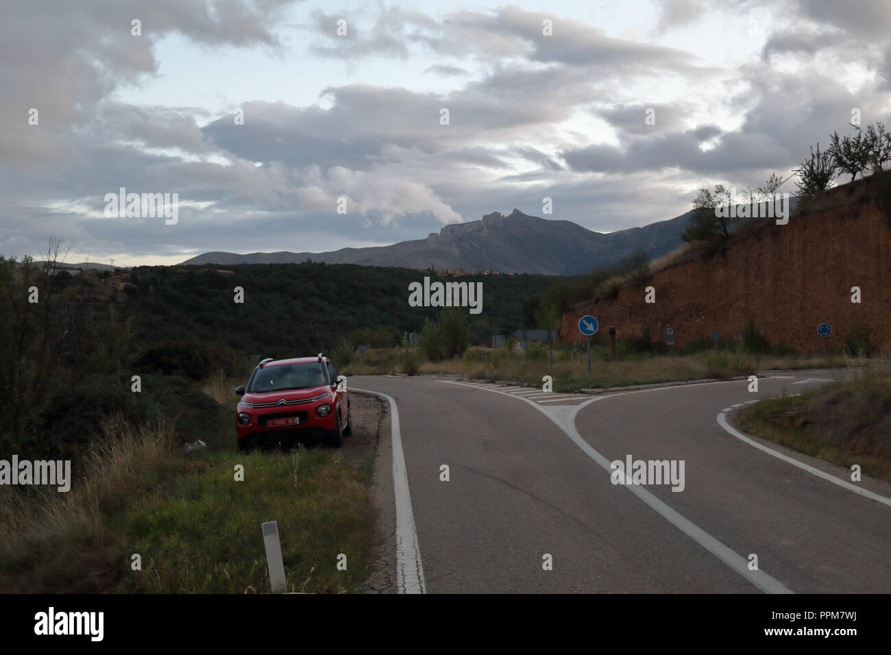 Un paesaggio di Aragona, Spagna, NUVOLOSO TRAMONTO in autunno. Un viaggiatore in auto rossa è parcheggiato a fianco di una rotatoria (sinistra-guida) durante un viaggio su strada Foto Stock