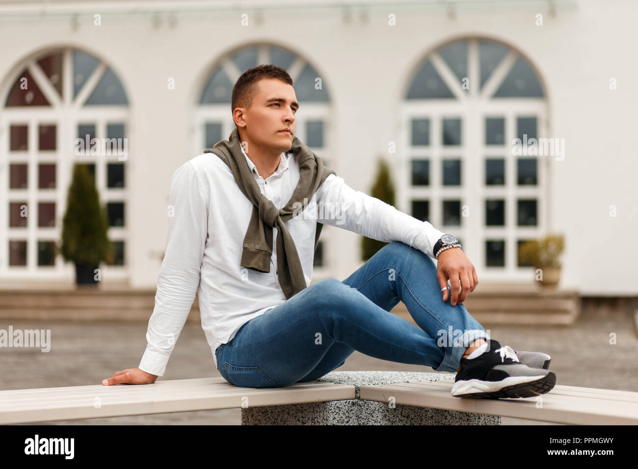 Elegante bel giovane uomo in camicia bianca con jeans e maglione seduta sul  banco in strada Foto stock - Alamy