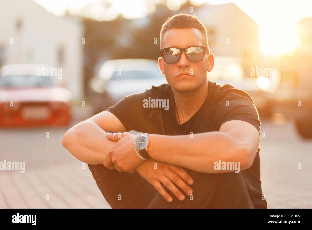 Elegante bel giovane con occhiali da sole nel quartiere alla moda di abiti neri si siede asfalto al tramonto Foto Stock