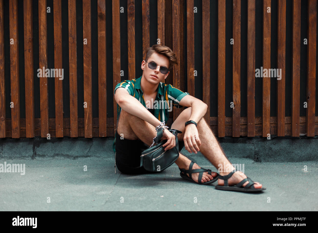 Moda giovane modello Uomo con occhiali da sole in una spiaggia shirt con pantaloncini nero seduto vicino a una parete in legno Foto Stock