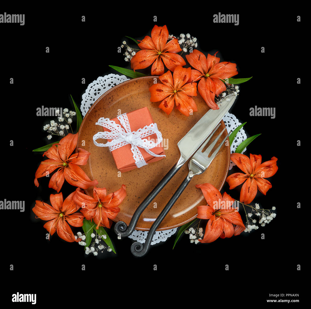 Piastra in terracotta decorata con gigli arancio fiori, confezione regalo legato con il bianco in pizzo e nastro esclusiva posate forgiato isolato su un nero backgroun Foto Stock