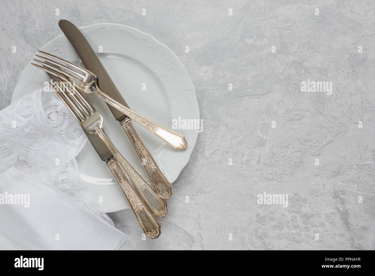 Posate in argento su una piastra di porcellana e tovagliolo bianco con merletto belga sono su sfondo grigio della superficie in calcestruzzo, con copy-spazio Foto Stock