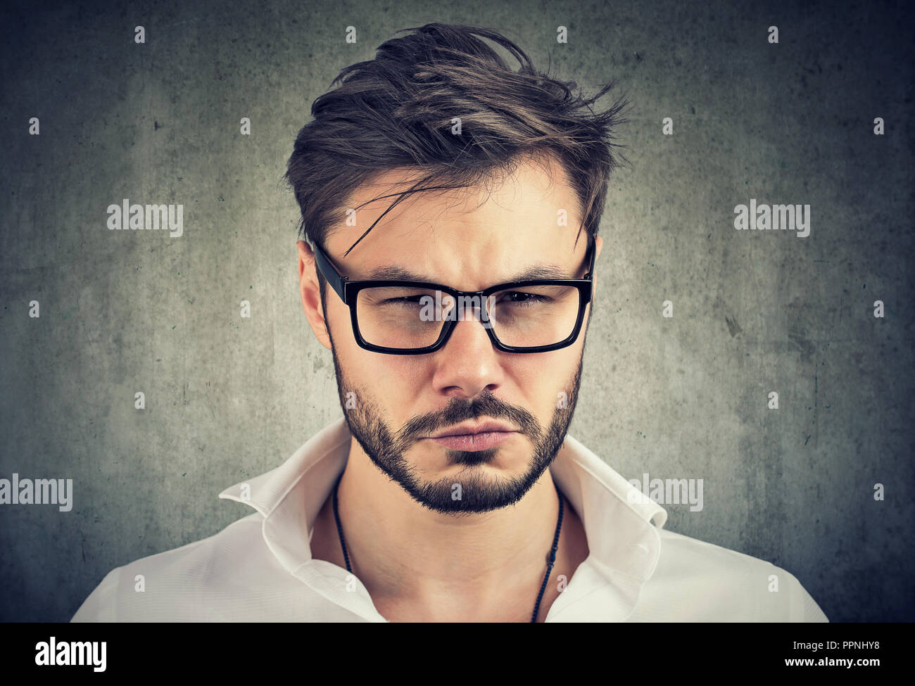 Giovane uomo barbuto in vetri cercando di gelosia e di sospetto in telecamera su uno sfondo grigio Foto Stock