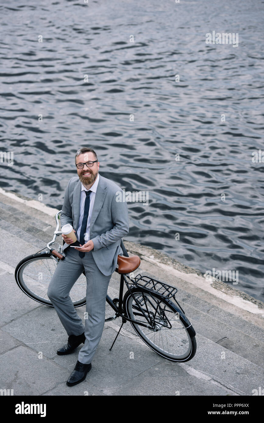 Imprenditore con caffè di andare e lo smartphone in piedi con la bicicletta sul quay Foto Stock