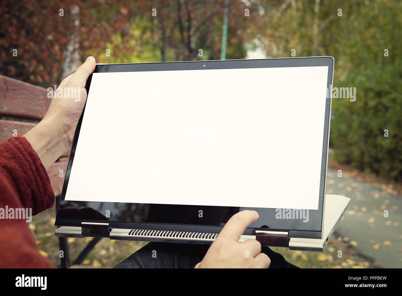 Uomo informale seduta su una panchina nel parco in autunno con le gambe incrociate e presentando il suo portatile con il bianco schermo vuoto. Toccare lo schermo sul computer portatile Foto Stock
