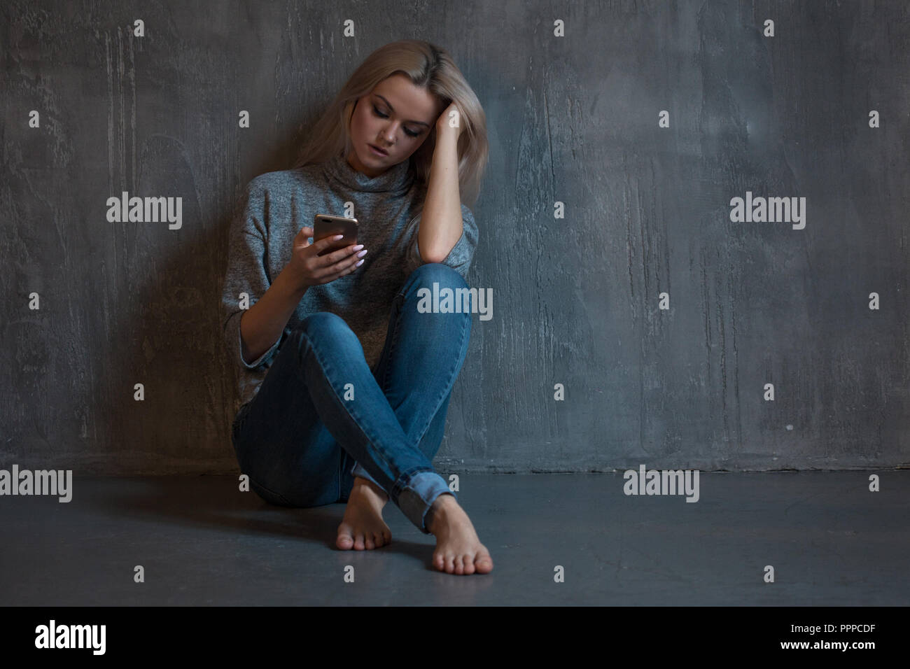 Helpline, assistenza psicologica. La sofferenza giovane donna seduta in un angolo con un telefono in mano. salute mentale, lo sfondo grigio Foto Stock