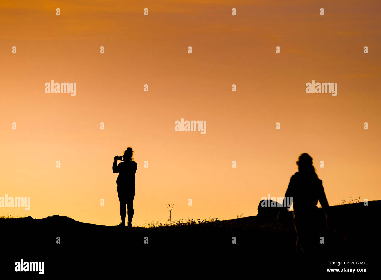 La silhouette di una figura utilizzando uno smartphone per fotografare un intenso tramonto su East pentire a Newquay in Cornovaglia. Foto Stock