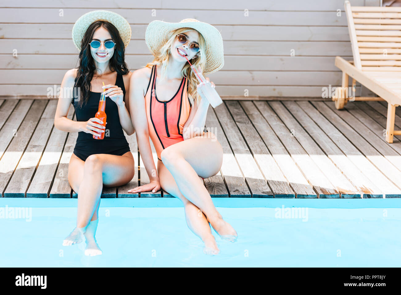 Bella giovani donne in costume da bagno a bere bevande in estate e sorridente in telecamera vicino alla piscina Foto Stock