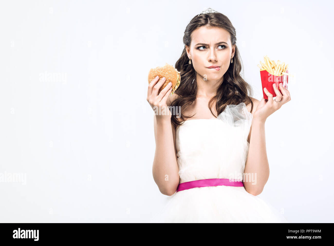 Considerato giovane sposa in abito da sposa con hamburger e patatine fritte cercando isolato su bianco Foto Stock