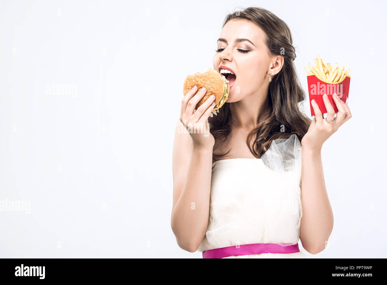 Hangry giovane sposa in abito da sposa di mangiare hamburger e patatine fritte isolato su bianco Foto Stock
