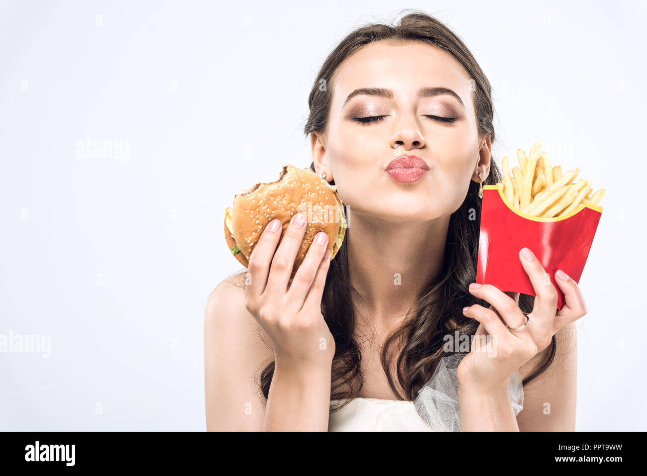 Giovane sposa in abito da sposa con hamburger e patatine fritte invio di kiss in telecamera isolato su bianco Foto Stock