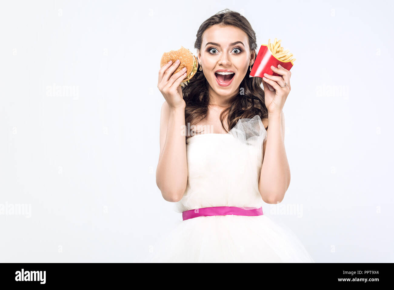 Scioccato giovane sposa in abito da sposa con hamburger e patatine fritte guardando la telecamera isolato su bianco Foto Stock