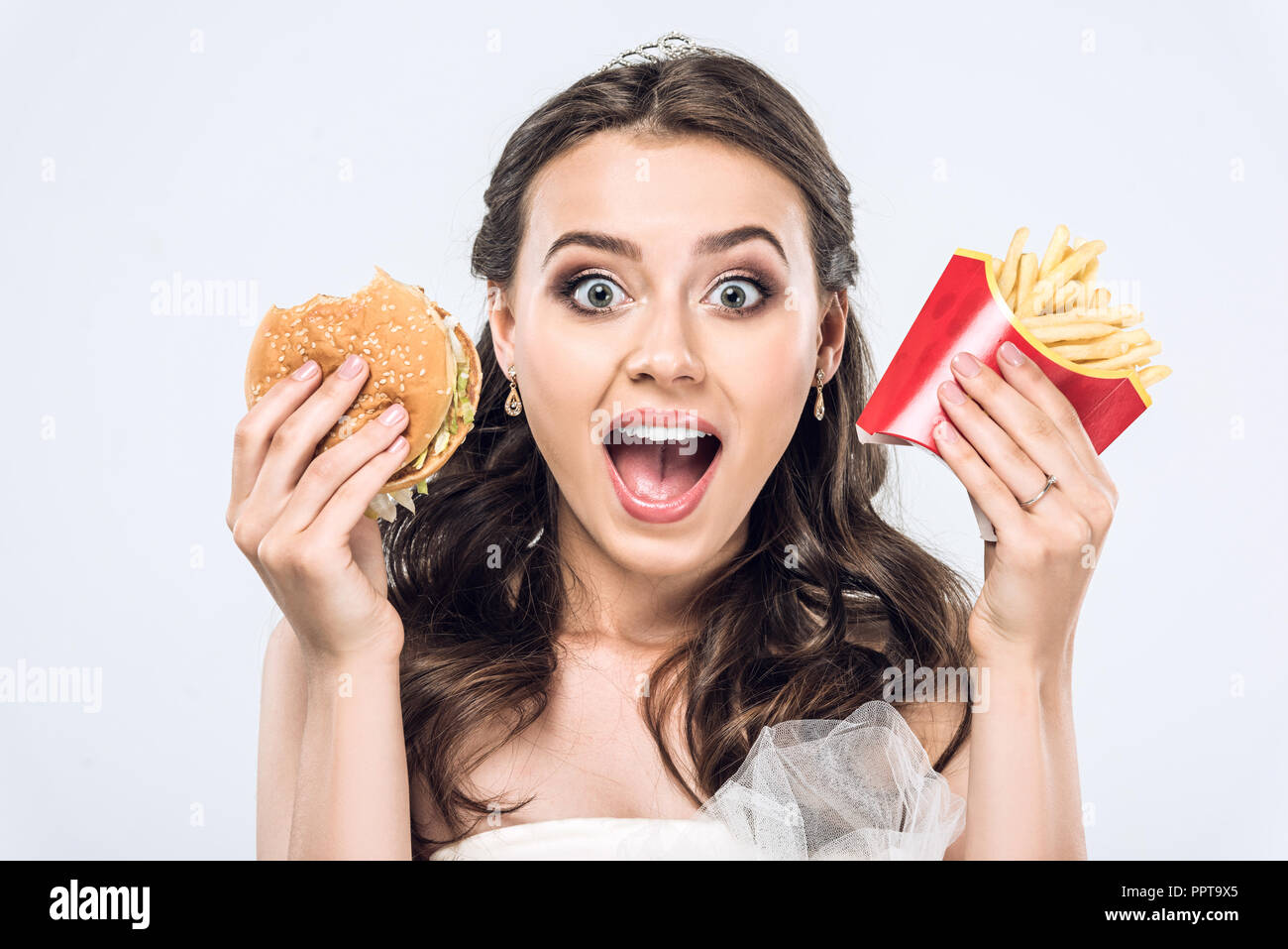 Close-up ritratto di scioccato giovane sposa in abito da sposa con hamburger e patatine fritte guardando la telecamera isolato su bianco Foto Stock