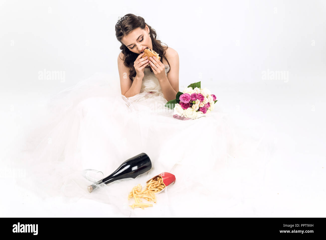 Angolo di alta vista del giovane sposa in abito da sposa di mangiare fast food mentre è seduto sul pavimento su bianco Foto Stock