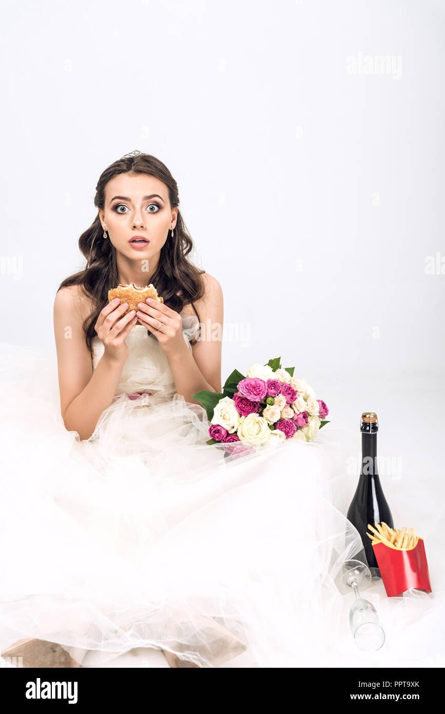 Scioccato giovane sposa in abito da sposa seduto sul pavimento con burger in mano su bianco Foto Stock