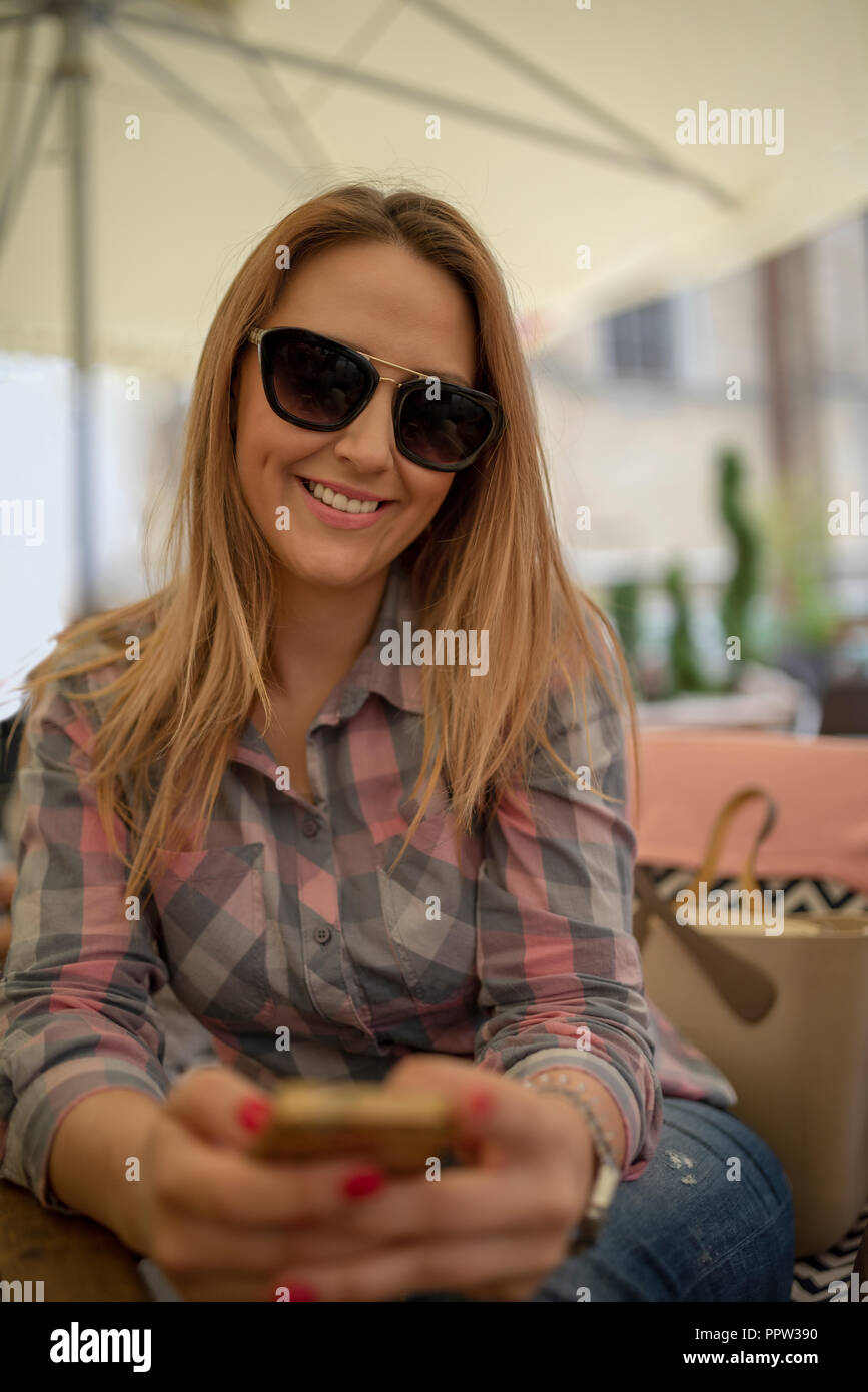 Affascinante ragazza giovane con telefono cellulare durante il riposo nella caffetteria. Bella femmina caucasica è texting messaggio sul telefono mentre vi rilassate in un cafè durante fr Foto Stock