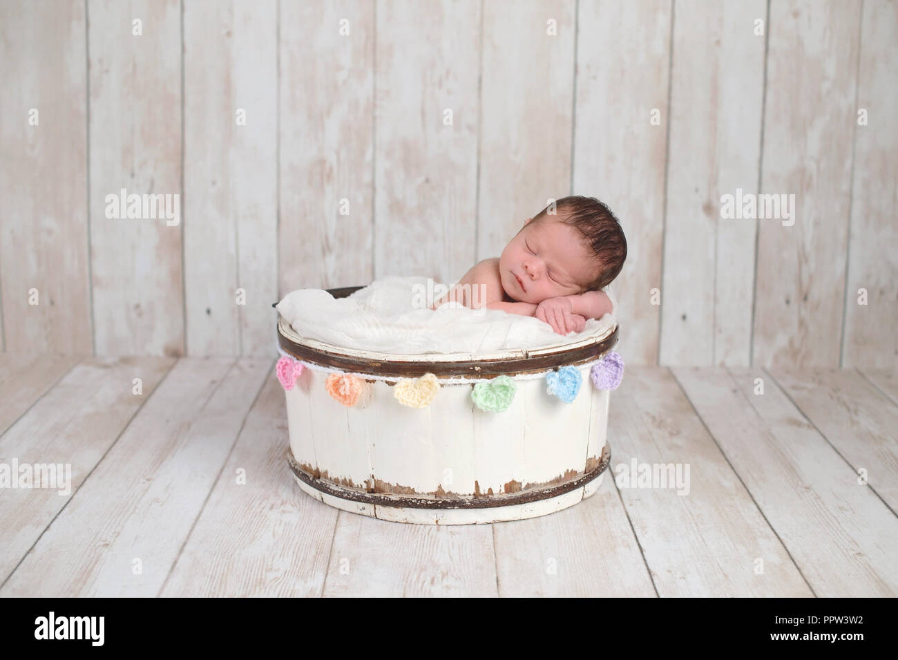 A due settimane di età neonato ragazza dorme in un piccolo, secchio di legno con un arcobaleno colorato ghirlanda di cuore. Girato in studio su un bianco, backg in legno Foto Stock