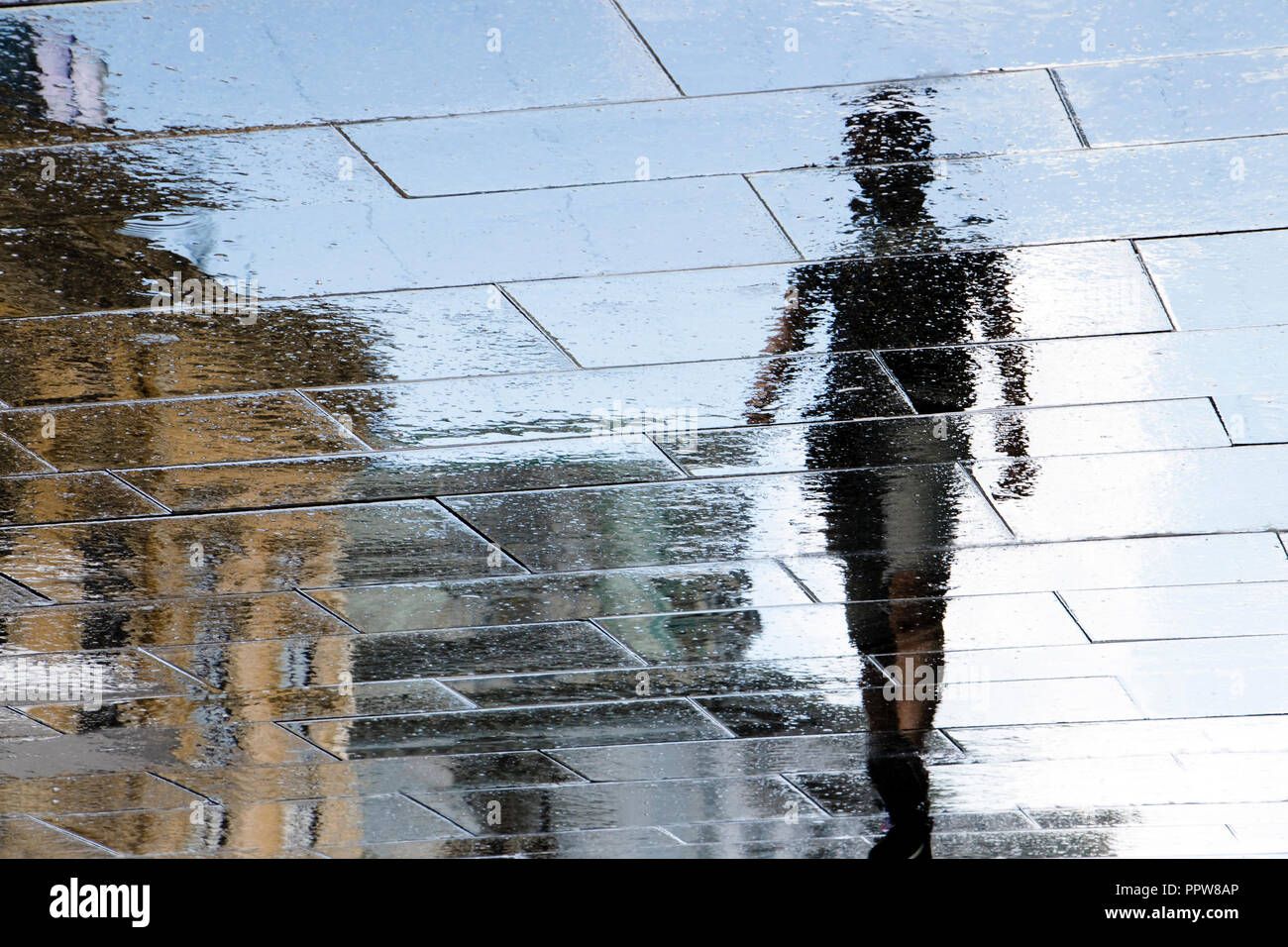 Sfocata riflessione silhouette di ombra in una pozza di un giovane uomo che cammina sulla città piovosa street dopo la pioggia Foto Stock