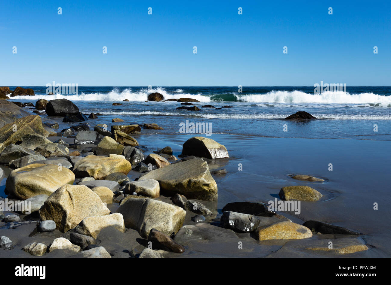 La mattina presto girato sulla spiaggia con la bassa marea, massi e sabbia in primo piano, di onde che si infrangono nella distanza contro un cielo blu senza nuvole. Foto Stock