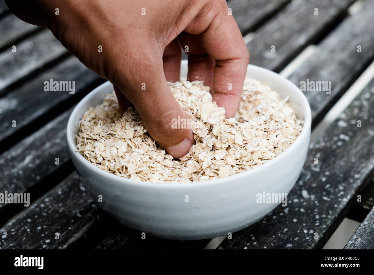 Primo piano della mano di un giovane uomo caucasico grabbing alcuni fiocchi d'avena da un bianco vaso in ceramica su tavola in legno rustico Foto Stock