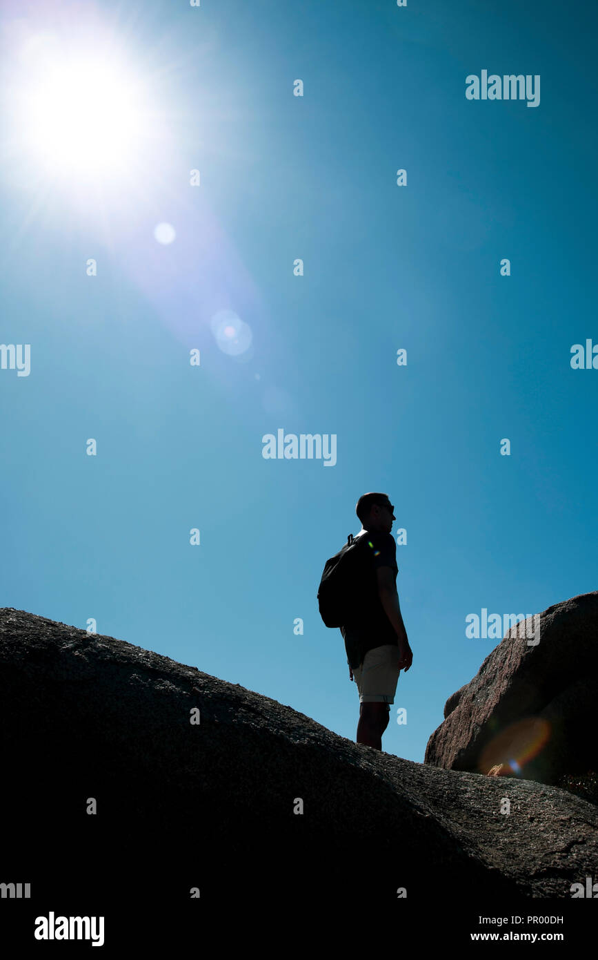 Primo piano di un giovane uomo caucasico portando uno zaino, nella parte superiore di una formazione di roccia, in Sardegna, Italia, retroilluminato con una sunbeam sulla sinistra Foto Stock