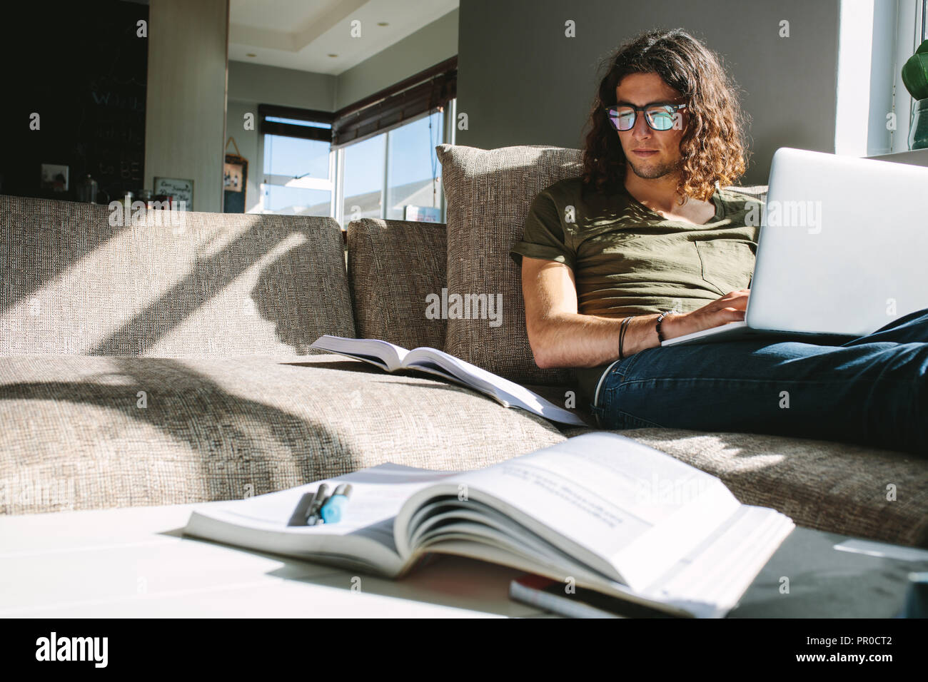 Giovane uomo studiando per gli esami seduti sul divano di casa con la luce del sole proveniente dalla finestra. Studente per esame su laptop con libri di fronte. Foto Stock