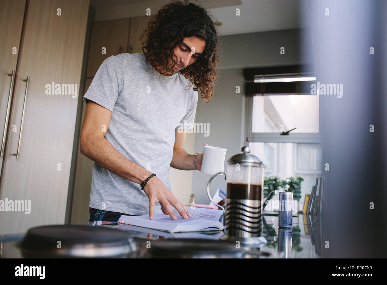 Studente la lettura di un libro di cucina mentre si beve il caffè. Sorridente studente per l'esame tenendo una tazza di caffè in piedi in cucina. Foto Stock
