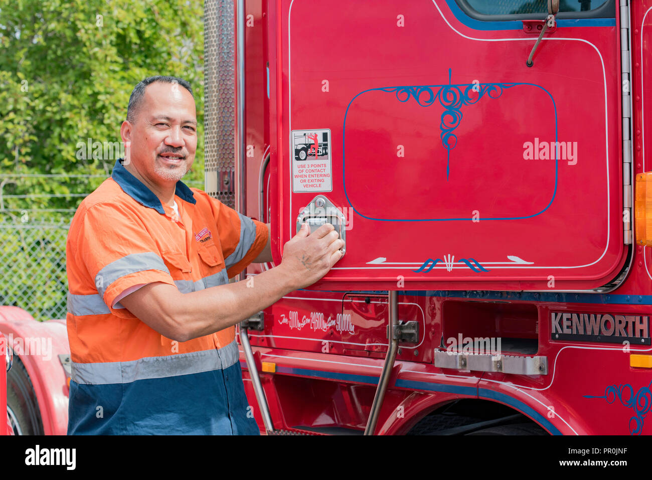 Un felice il samoano camionista in indumenti ad alta visibilità, arrampicata in una cabina di un rosso semi rimorchio prime mover carrello Foto Stock