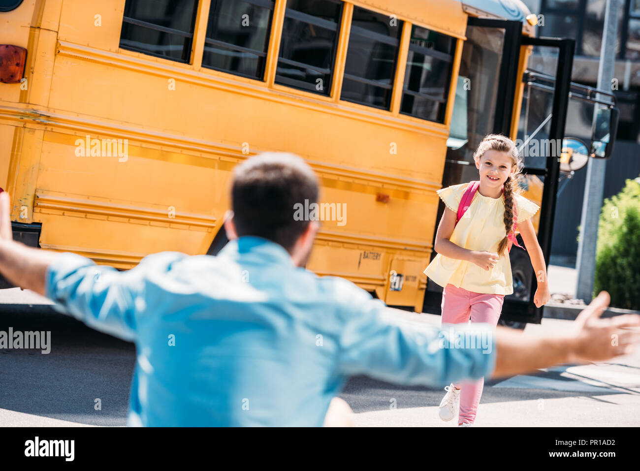 Vista posteriore del padre con le braccia aperte in attesa di felice piccola figlia mentre era in esecuzione dal bus di scuola Foto Stock