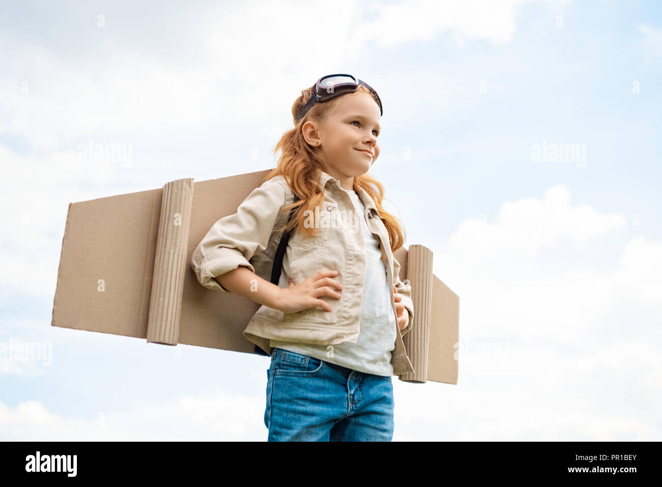 Basso angolo vista del bambino con il piano della carta delle ali sulla schiena e occhiali di protezione sulla testa contro blu cielo molto nuvoloso Foto Stock