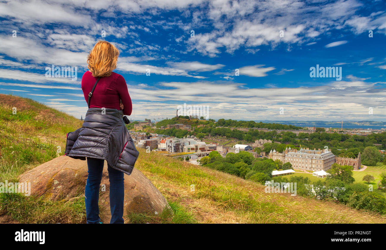 La donna caucasica guardando fuori su Edimburgo dalle falesie di Holyrood Park su una luminosa giornata soleggiata con un cielo blu e nuvole bianche Foto Stock