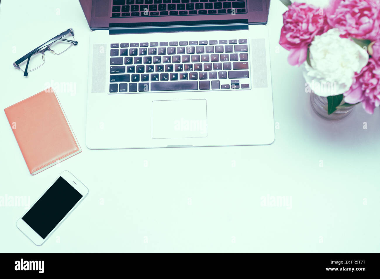 Il luogo di lavoro con computer portatile, bicchieri, notebook e bianco e rosa peonia fiori sul tavolo bianco sullo sfondo. Piatto, laici vista superiore Foto Stock