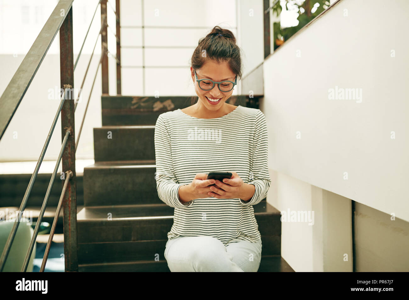 Giovane imprenditrice asiatica con gli occhiali e sorridente seduti sulle scale in un ufficio utilizzando un cellulare durante la sua rottura Foto Stock