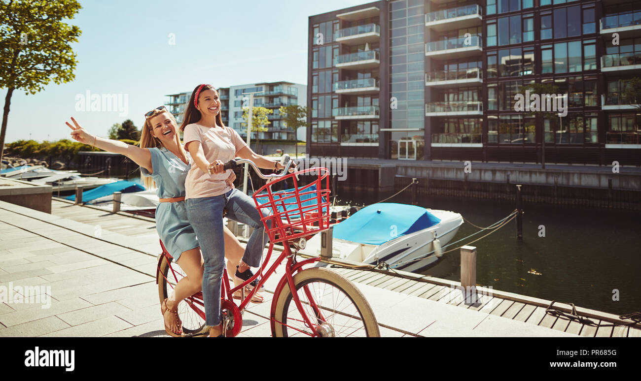 Due giovani spensierati fidanzate di ridere mentre in sella a una moto insieme lungo la passeggiata del lungomare in estate Foto Stock