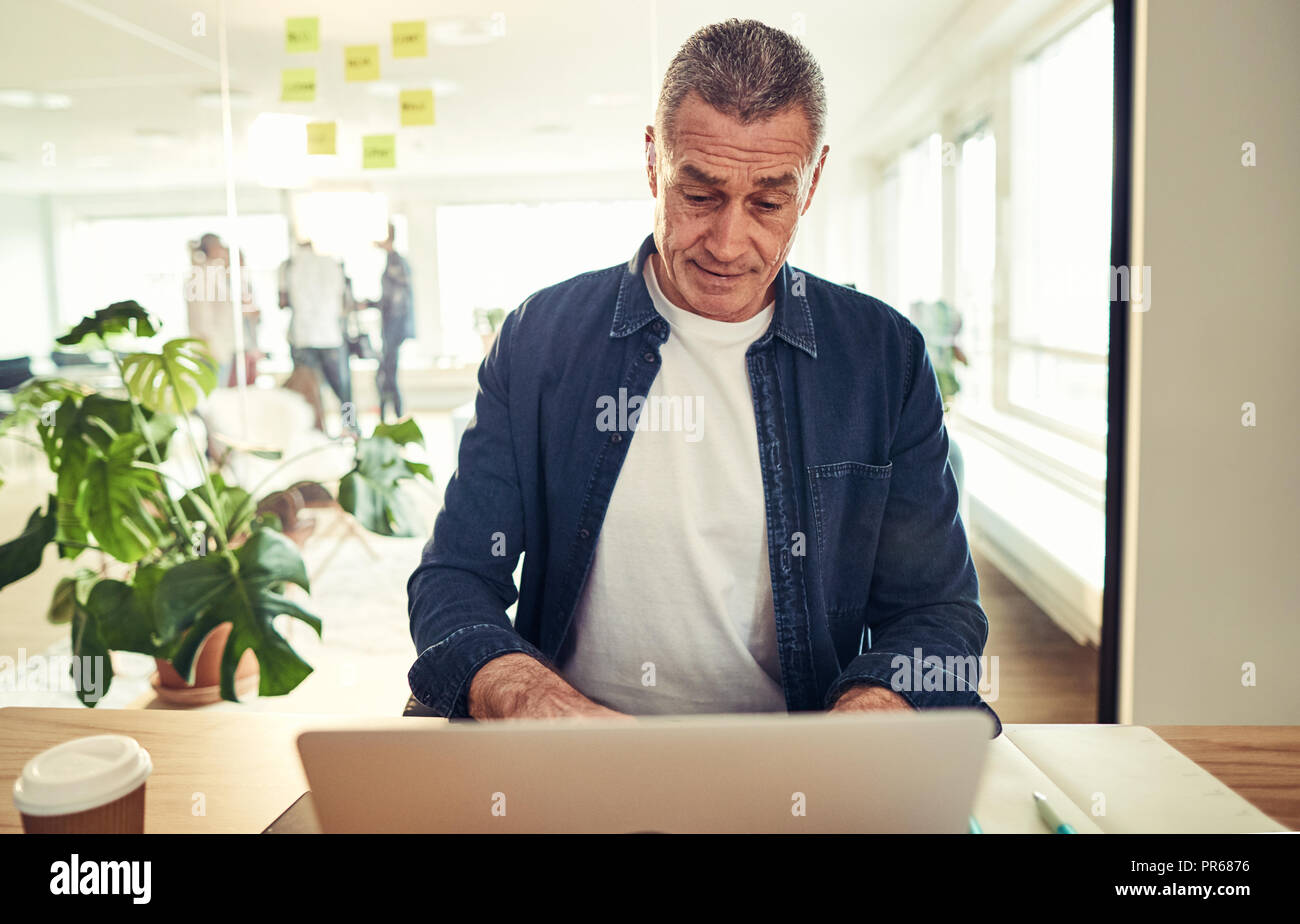 Casualmente vestito imprenditore maturo seduti ad una scrivania nel suo ufficio in un duro lavoro su un notebook Foto Stock
