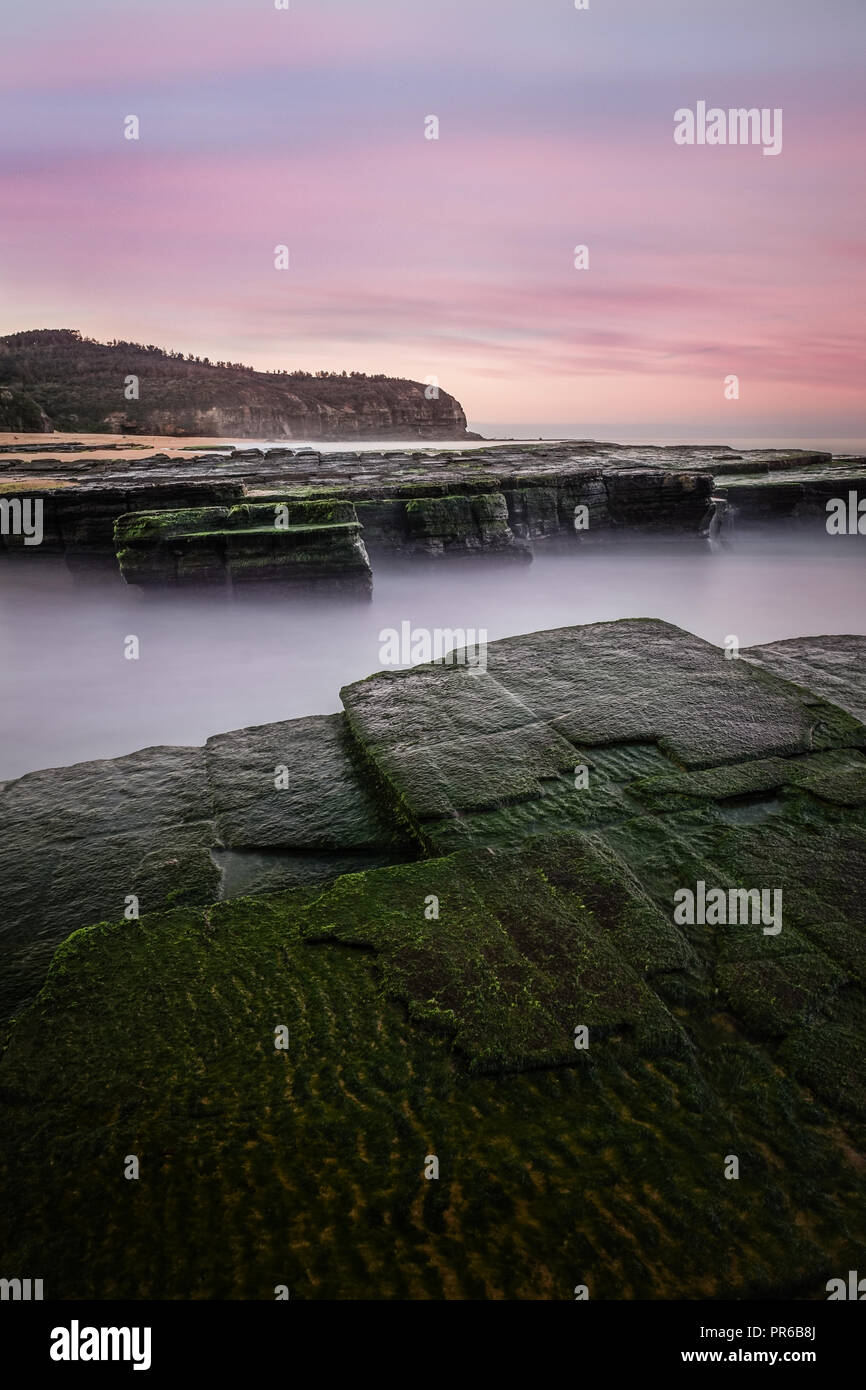 Rosa luminoso Cielo di tramonto con rocce di muschio in primo piano dalla spiaggia Turimetta. Foto Stock
