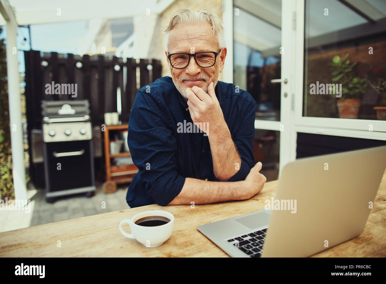 Sorridente uomo senior gustando una tazza di caffè e di lavorare su un laptop mentre si siede a un tavolo esterno sul suo patio Foto Stock